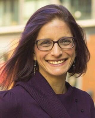 Portrait of a caucasian woman with short, chin-length dark brown and dark purple hair. She has purple square glasses on and is wearing a dark purple sweater. She faces the camera, smiling.